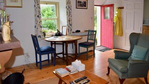 Een gezellige woonkamer met een houten vloer, een ronde eettafel, twee blauwe stoelen, een groene fauteuil en een kleine boekenkast.