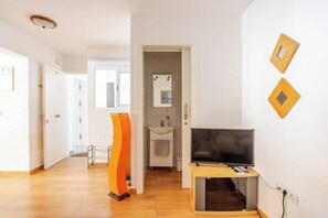 A modern living room with a television on a wooden stand, a white wall with two decorative wooden frames, and a hallway leading to a bathroom.