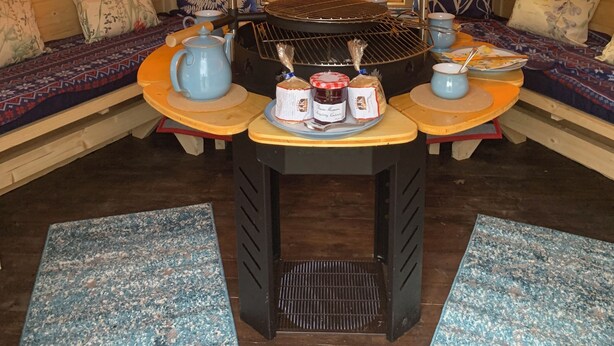 Un interior acogedor con techo de madera, una mesa central con parrilla y asientos con cojines estampados.