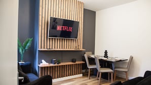 A modern living room with a flat-screen TV mounted on a wooden panel wall, a dining area with a table and chairs, and a sofa. - Entire Modern Home in Manchester For You (Manchester)