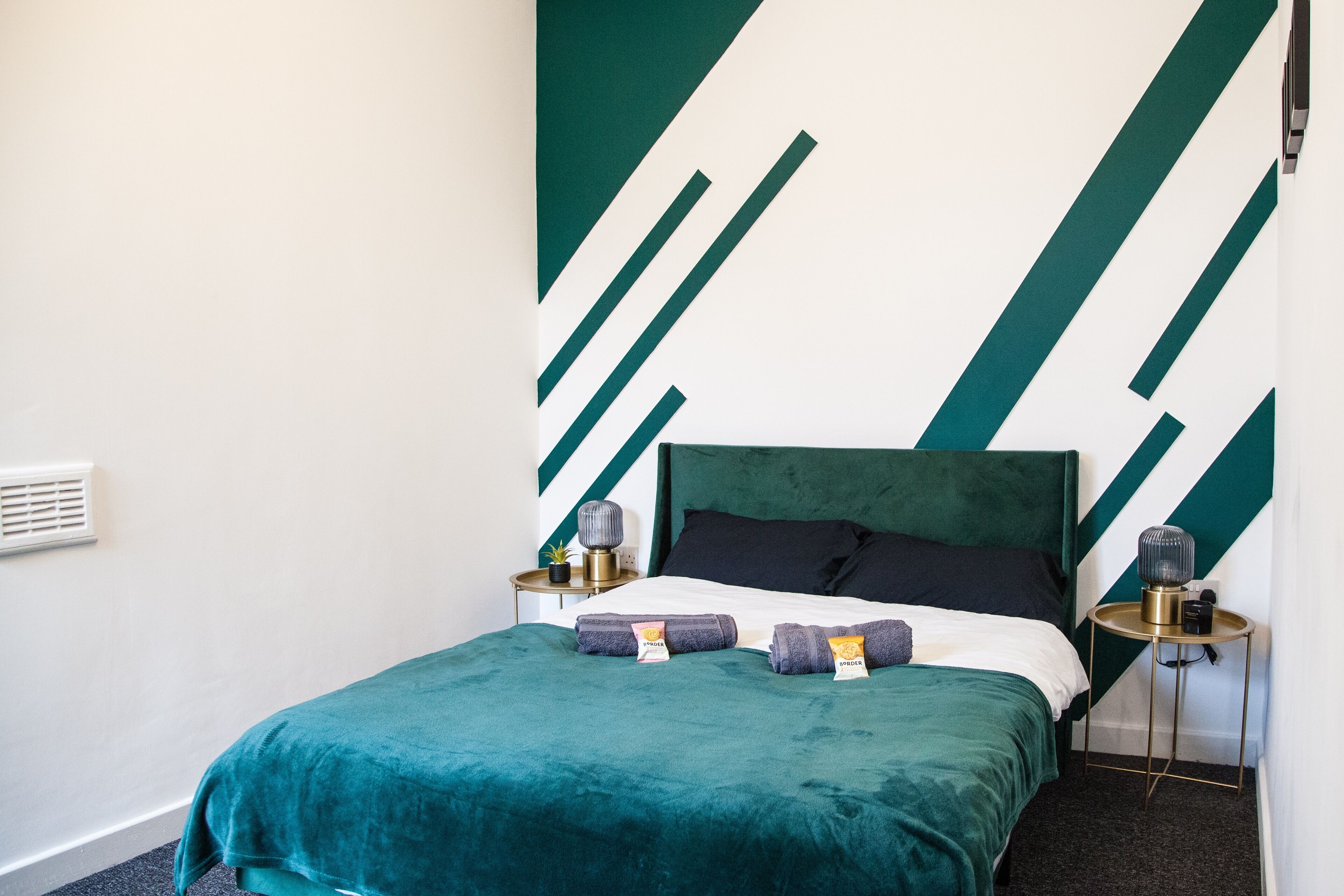 A neatly made bed with a teal bedspread and pillows, two bedside tables with lamps, and a wall with a geometric pattern.