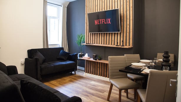 A modern living room with a black sofa, a dining table set for four, and a mounted TV displaying "NETFLIX". - Entire Modern Home in Manchester For You (Manchester)