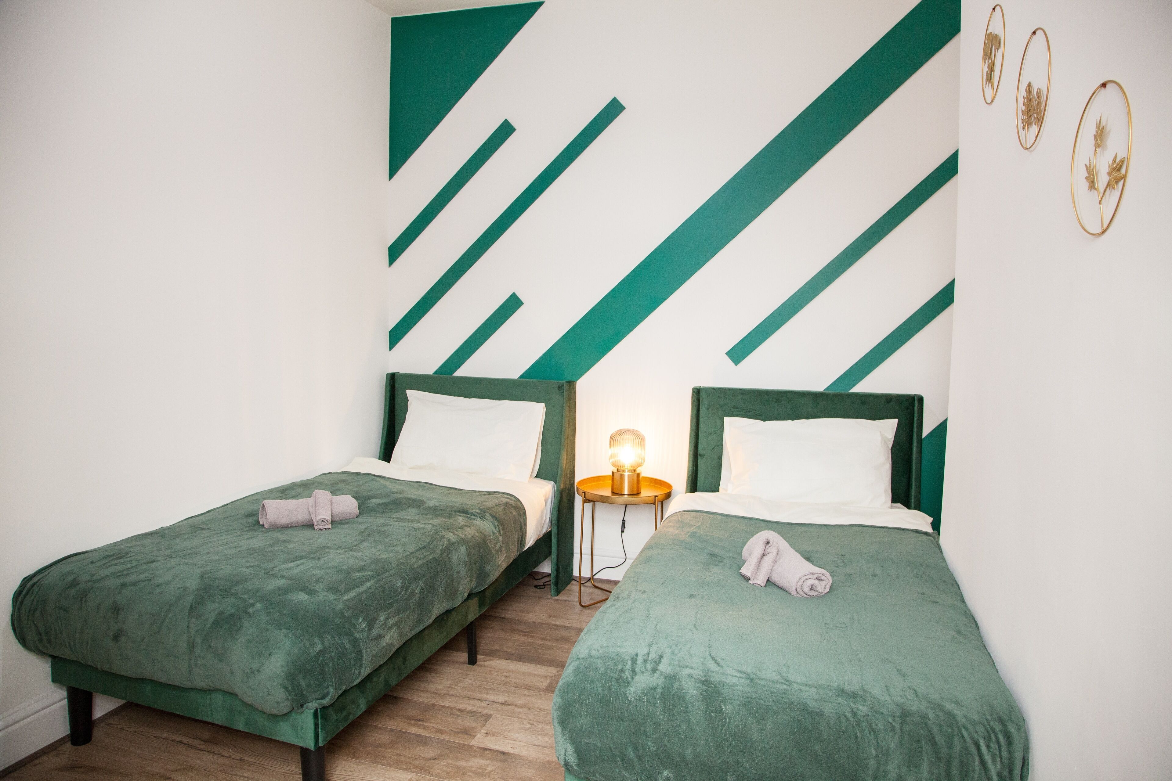 Duas camas de solteiro com roupa de cama verde, uma pequena mesa de cabeceira com abajur, e uma parede com padrões geométricos verdes e brancos.
