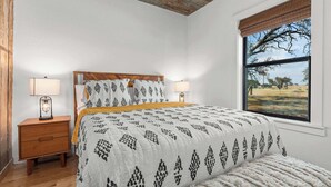 A bedroom with a bed, two bedside tables with lamps, a window with a view of trees, and a rustic ceiling. - Starry Night Cottage 1 (Stonewall)