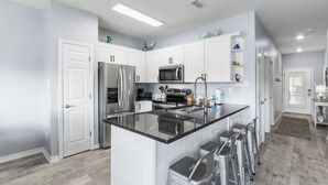 A modern kitchen with white cabinetry, stainless steel appliances, and a central island with stools. - Romar Lakes 103B (Orange Beach)