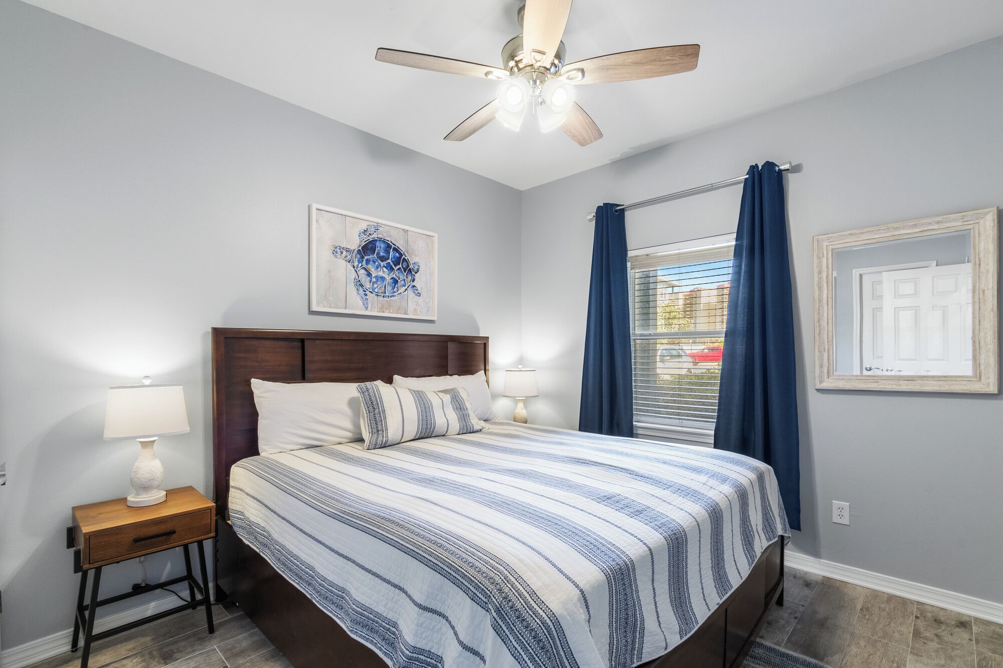 Une chambre à coucher comprenant un lit, une table de chevet, un ventilateur de plafond, une fenêtre avec des rideaux et un tableau accroché au mur.