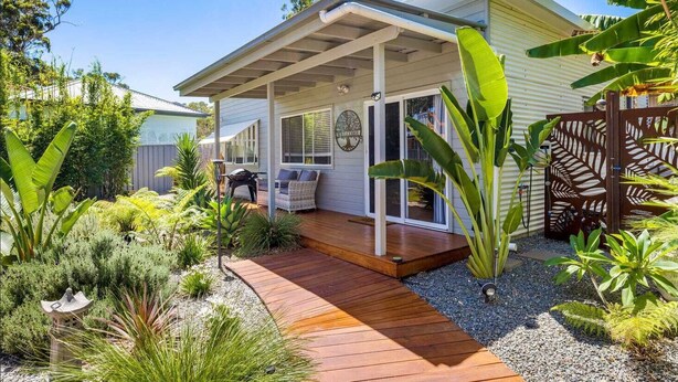 Una casa con porche cubierto, un pequeño jardín con plantas tropicales y una terraza de madera.