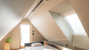 A modern loft bedroom with a sloped ceiling, a large skylight, a bed with a headboard, a potted plant, and a glass railing. - HOTEL NICE (Laupheim)