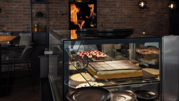 A bakery display case with various desserts, a brick wall, and a hanging light fixture. - HOTEL NICE (Laupheim)
