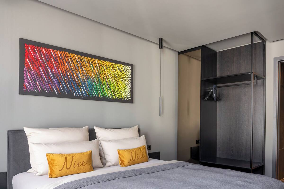 Un dormitorio moderno con una cama grande, dos almohadas con la palabra "Nice" y un cuadro abstracto colorido en la pared, además de un vestidor.