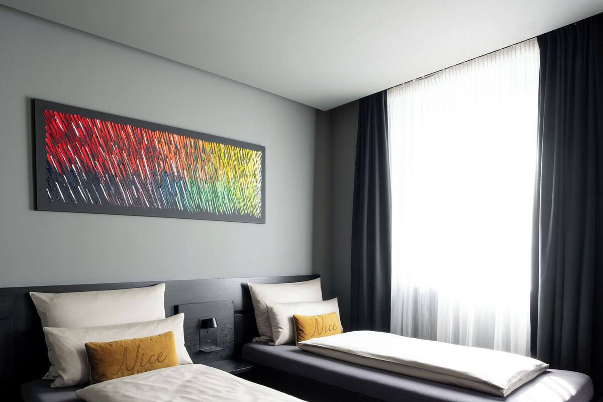 Una habitación de hotel moderna con dos camas, un cuadro abstracto grande y vistas de la ciudad a través de cortinas translúcidas.