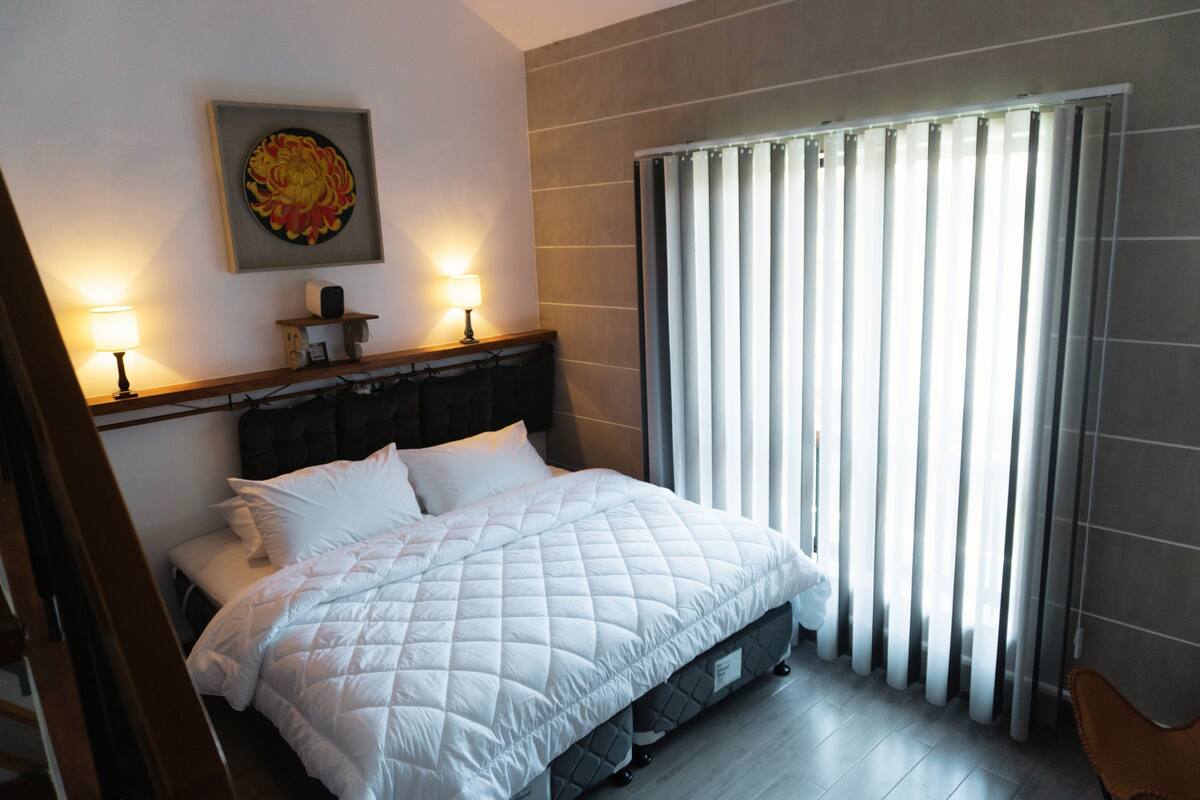 Una cama bien hecha con un edredón blanco acolchado, dos lámparas de noche, una obra de arte enmarcada sobre la cama y una ventana con persianas verticales.