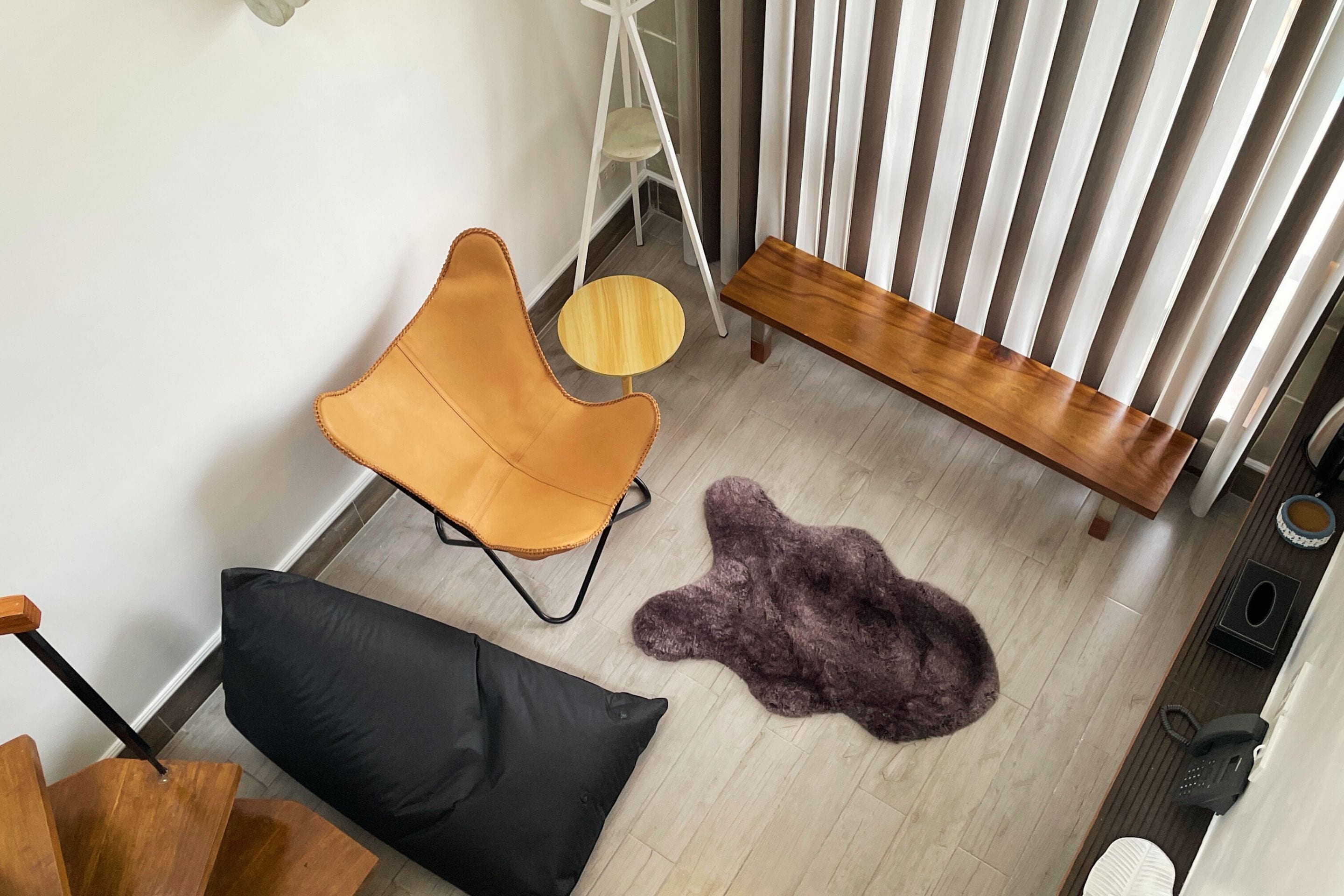 Una habitación con piso de madera, una silla negra, una silla amarilla, una pequeña mesa redonda, un banco de madera, un tapete mullido, un radiador blanco y una silla blanca con un cojín blanco.
