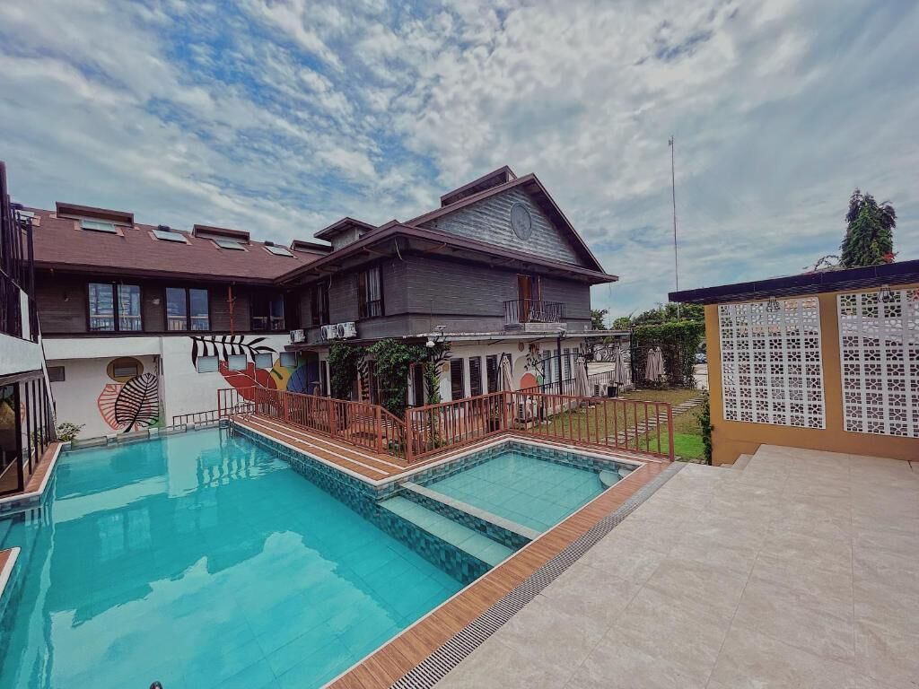 Une piscine avec une terrasse en bois, un patio couvert et un bâtiment doté d’un toit en pente et de grandes fenêtres.
