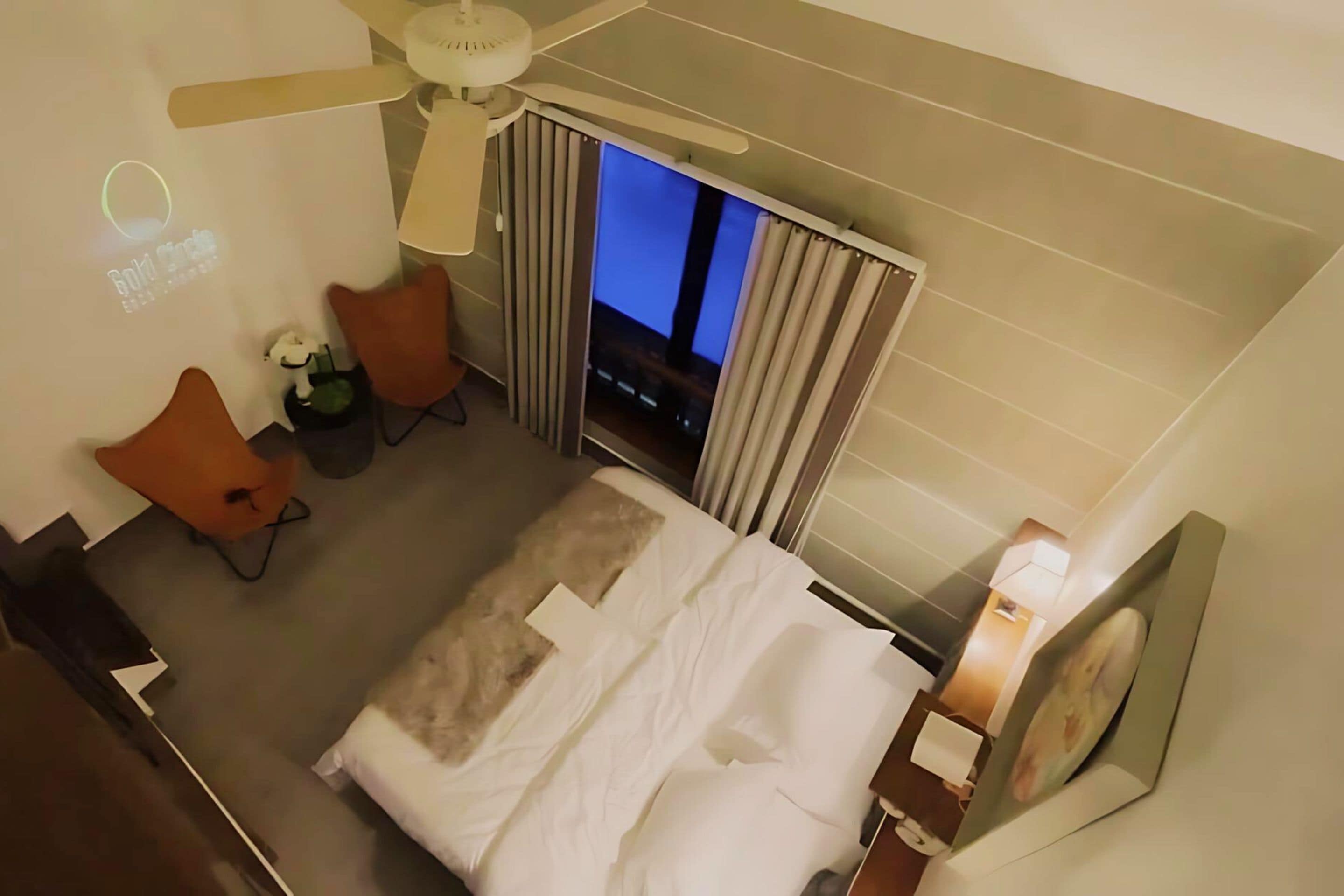 Ein Hotelzimmer mit Bett, Schreibtisch, Sessel, Fernseher und Deckenventilator.