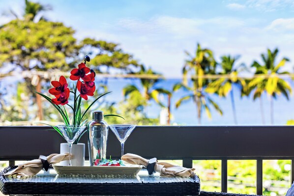 Una mesa con una copa de martini, una botella y un florero con flores rojas, ubicada en un balcón con vista a una playa tropical con palmeras.