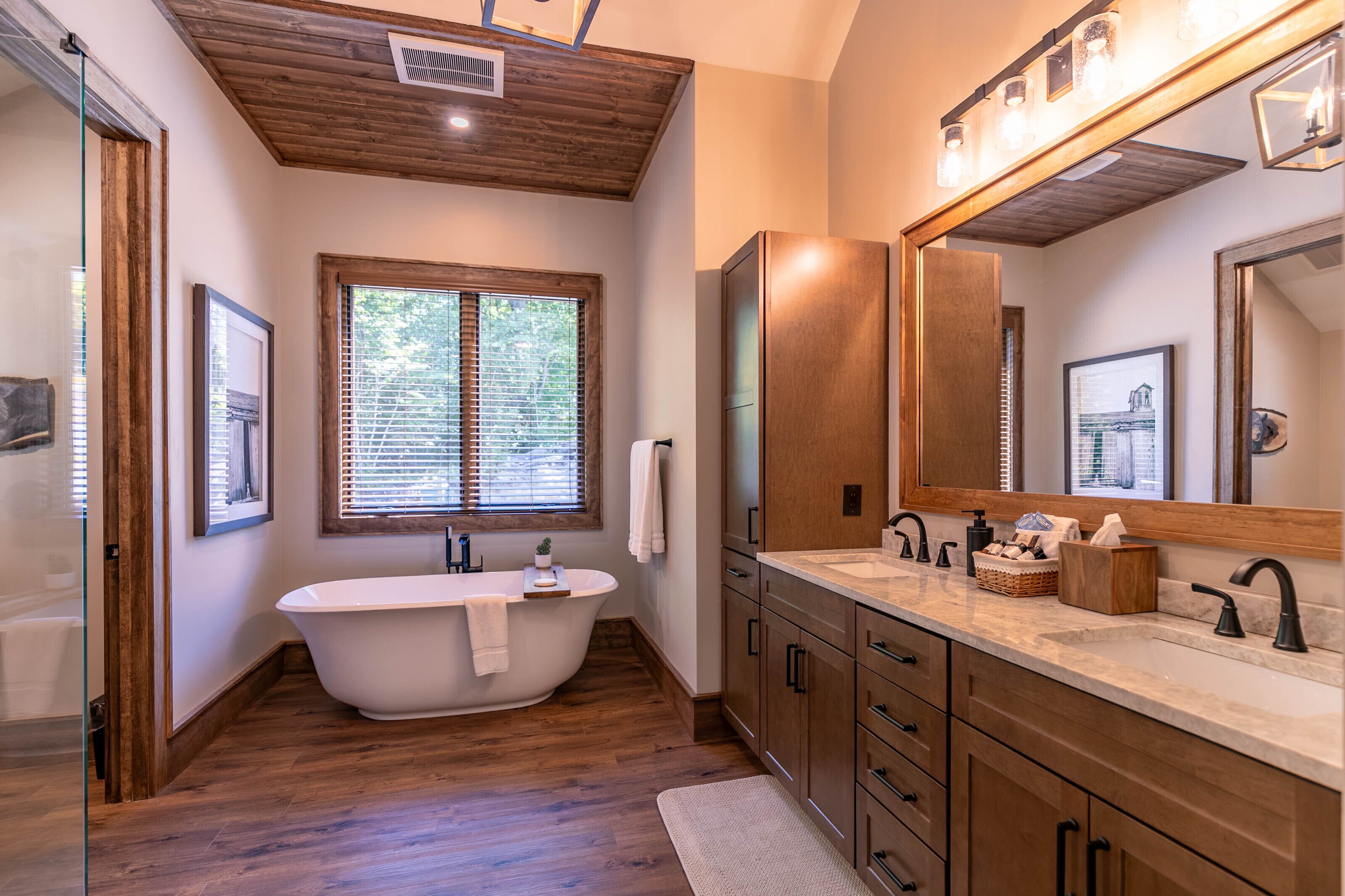 A modern bathroom with a freestanding bathtub, wooden cabinets, a large mirror, and a window with blinds.