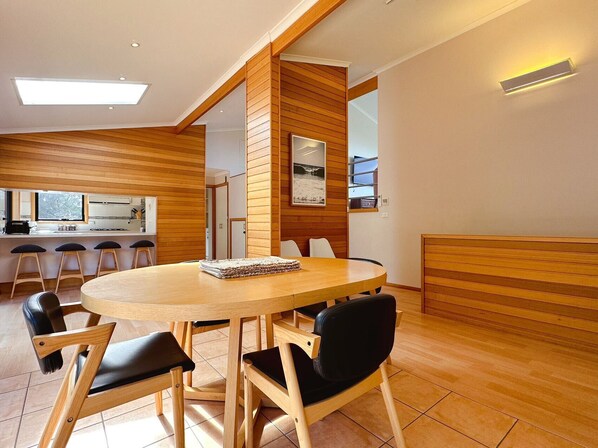 A modern dining area with a wooden table and chairs, a kitchen in the background, and a large window.