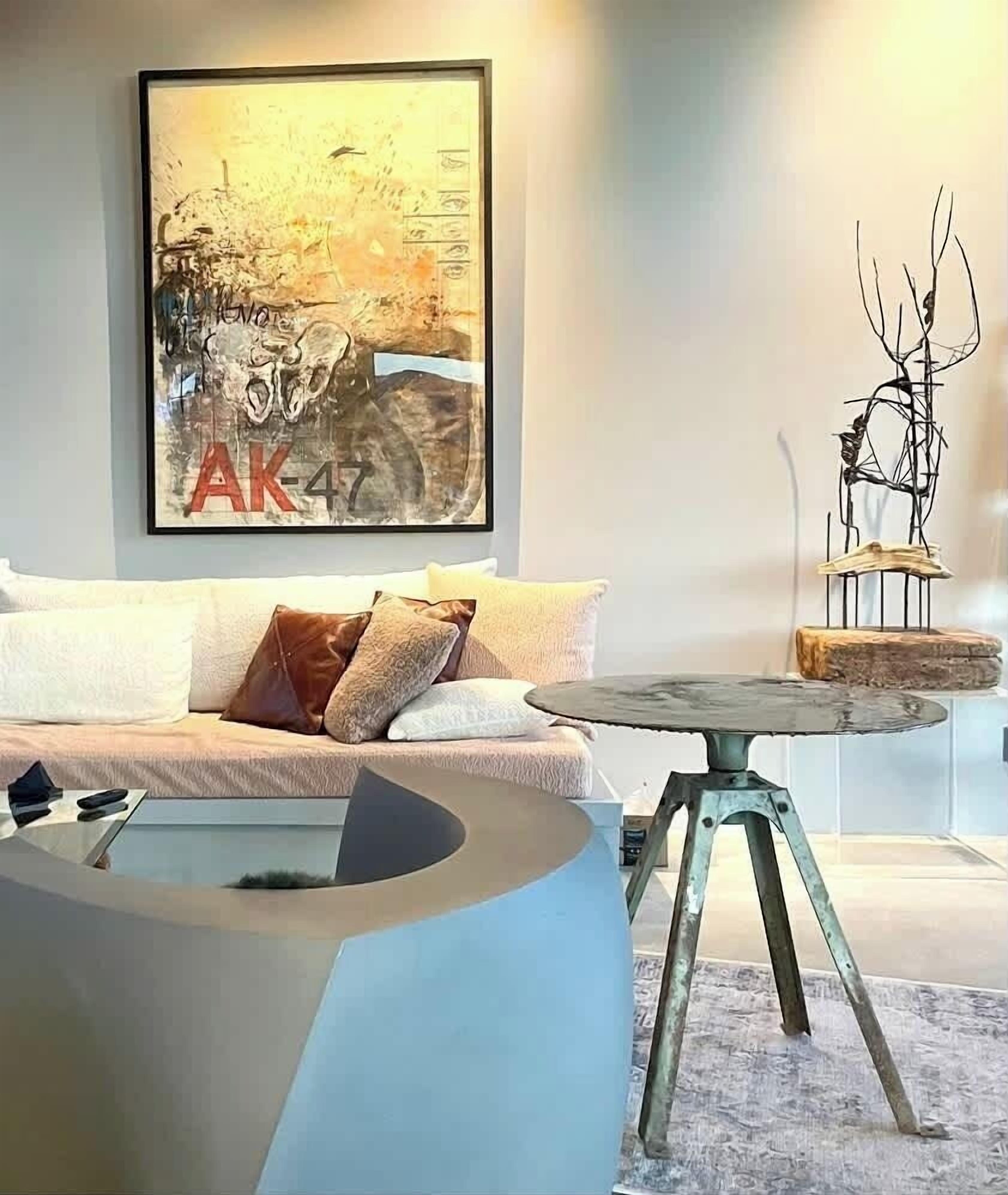 Ein modernes Wohnzimmer mit einem runden Couchtisch, einer Couch mit Kissen, einem gerahmten abstrakten Gemälde und einer Metallskulptur auf Holzfuß.