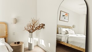 A bedroom with a bed, a round side table, a vase with branches, a mirror, and a wall-mounted picture.