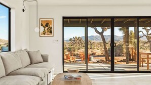 A modern living room with a beige sofa, a wooden coffee table with books, and a large sliding glass door leading to an outdoor patio with desert views.
