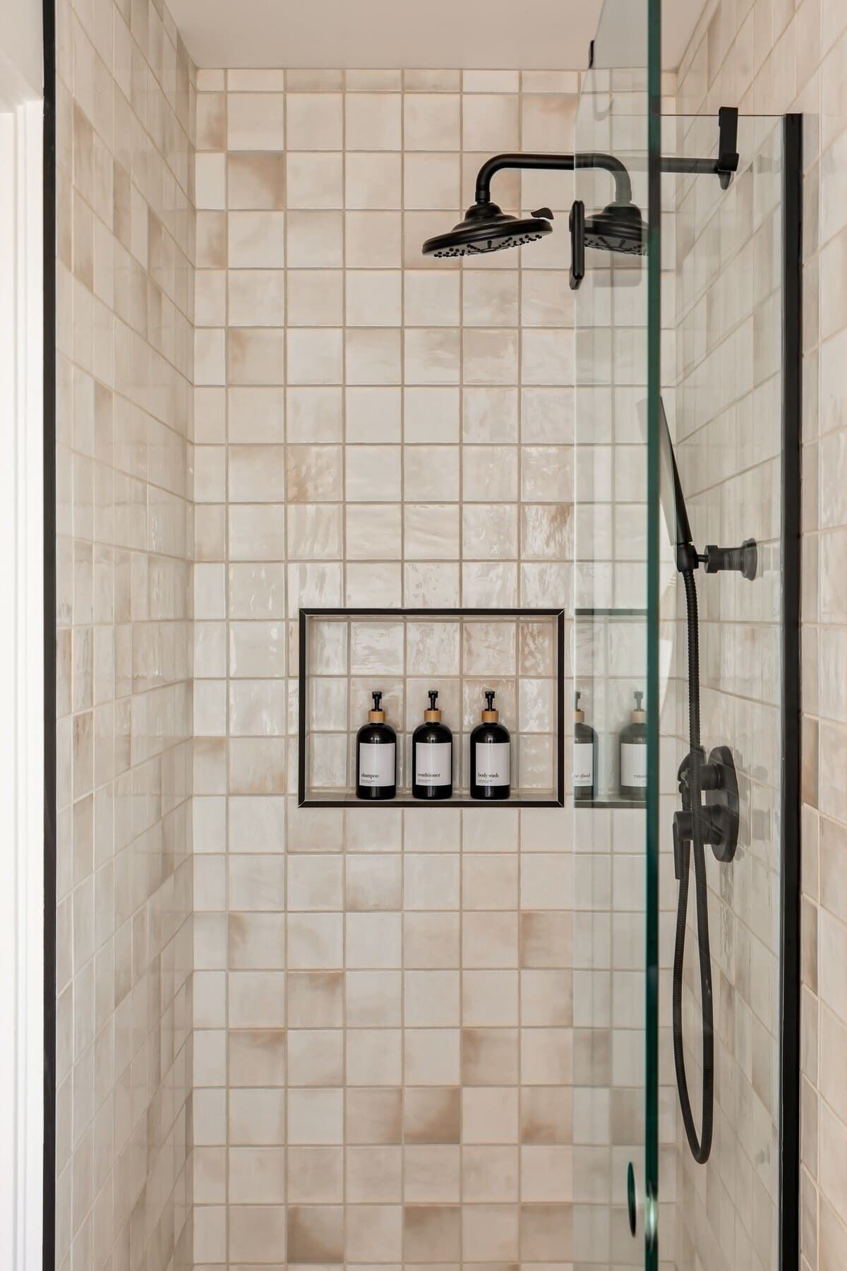 A glass-enclosed shower with beige tiles, a handheld showerhead, and a glass shelf with three bottles labeled "shampoo," "body wash," and "shampoo."