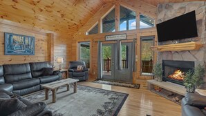 L’intérieur d’une cabane en rondins, avec une cheminée, des meubles en cuir, un téléviseur fixé au mur et une grande fenêtre.