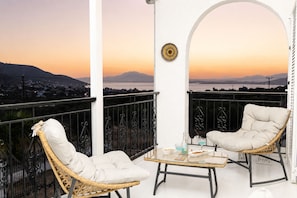 A balcony with two wicker chairs, a small table, and a view of a cityscape at sunset. - Villa Aelia Luxury Maisonette (ΑΙΑΝΤΕΙΟ ΣΑΛΑΜΙΝΑ)