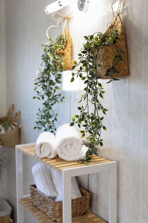 Une salle de bain avec une étagère blanche où sont posées des serviettes et un panier en osier, une plante suspendue et un miroir au mur.
