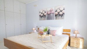 Quarto com cama de madeira, mesa de cabeceira com luminária, apoio para livros e prateleira na parede com uma planta.
