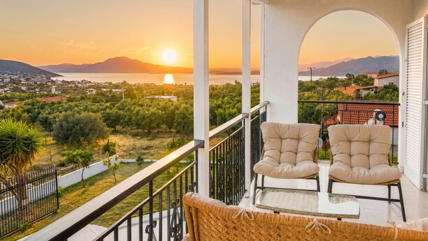 Uma varanda com duas cadeiras acolchoadas, uma mesa de vidro e vista para uma cidade costeira ao pôr do sol.