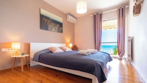 A bedroom with a bed, a nightstand, a lamp, an air conditioner, a window with curtains, and a view of the sea. - 4-bedroom villa in lovely Mili with AC, WiFi. Enjoy a relaxing stay (MILI)