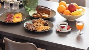 A dining table with plates of food, a bowl of fruit, a glass of orange juice, and a vase with a plant. - 4-bedroom villa in lovely Mili with AC, WiFi. Enjoy a relaxing stay (MILI)