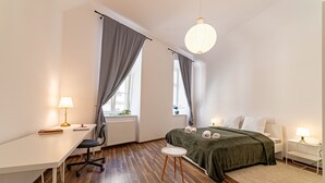 A modern bedroom with a wooden floor, a desk with a lamp, a bed with a green blanket, and a hanging light fixture. - RentPlanet - plac Uniwersytecki (Wroclaw)