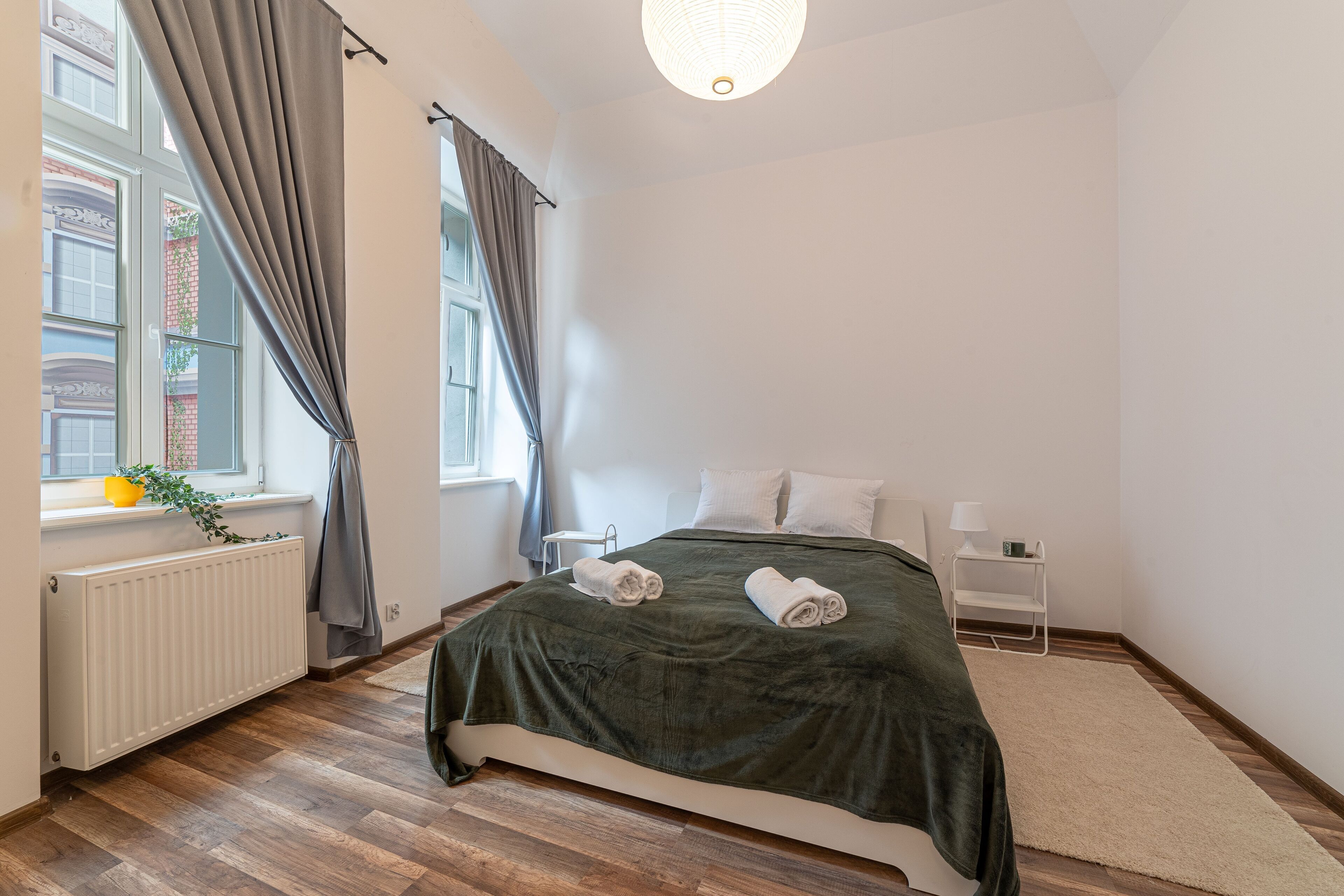 Ein ordentlich eingerichtetes Schlafzimmer mit einem großen Bett, einem Heizkörper, einem Fenster mit Vorhängen und einem Nachttisch mit Lampe.