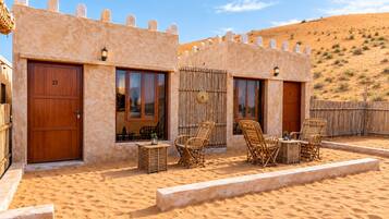A desert-style building with two doors, a small outdoor seating area with wicker furniture, and a sandy landscape in the background.