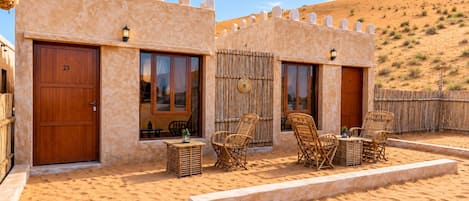 A desert-style building with two doors, a small outdoor seating area with wicker furniture, and a sandy landscape in the background.
