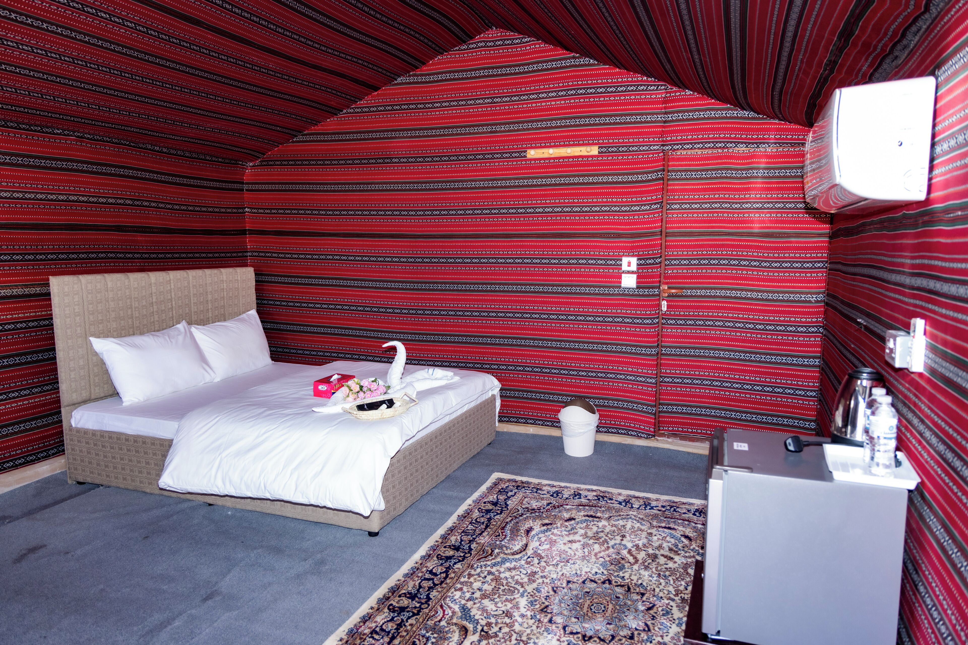 Een kamer met een bed, een klein tafeltje, een prullenbak en een wandlamp.