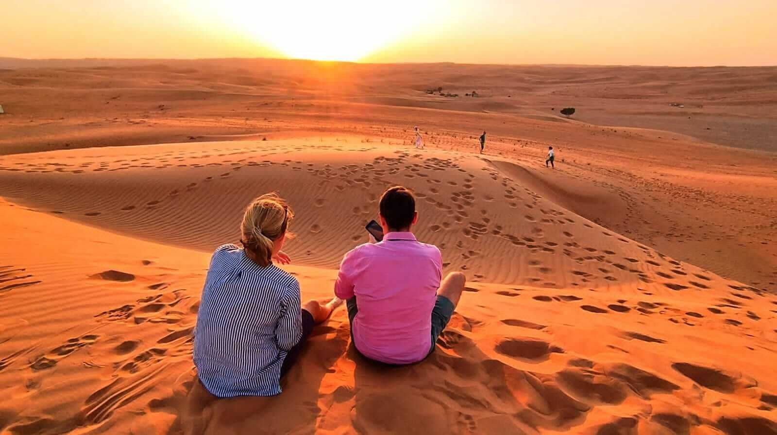 Dos personas sentadas en una duna de arena con un atardecer de fondo.