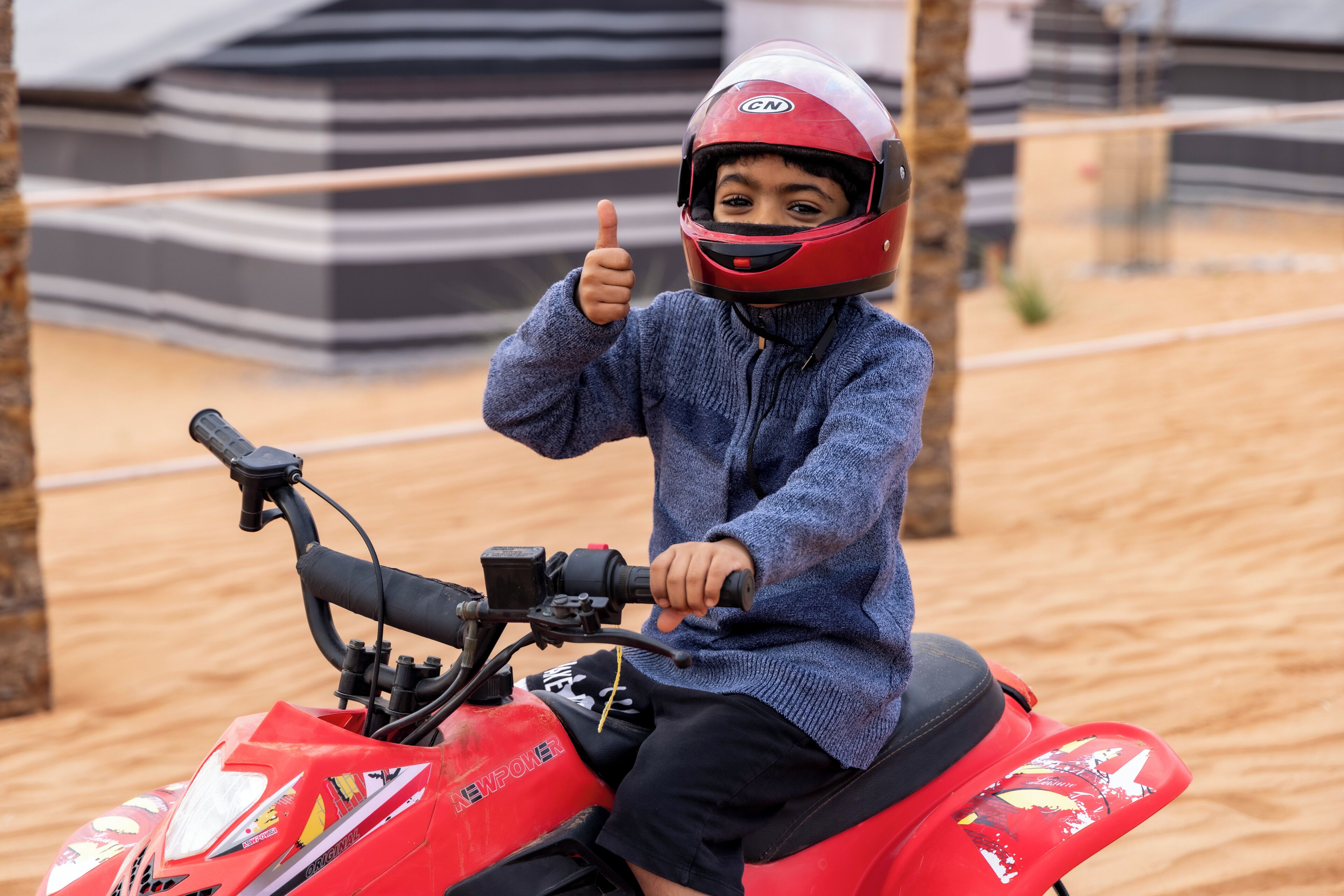 A child on a red ATV with a helmet, giving a thumbs-up.
