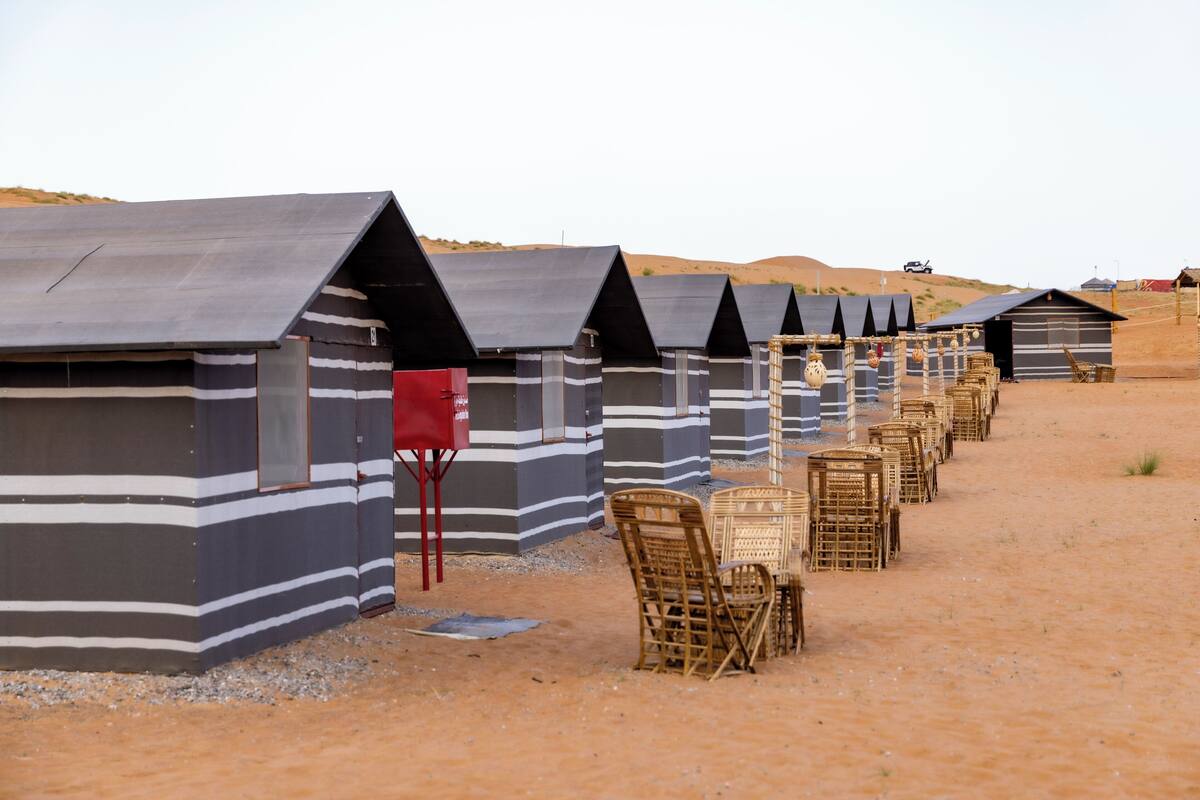 A row of striped beach huts with a sandy ground and a few wicker chairs.