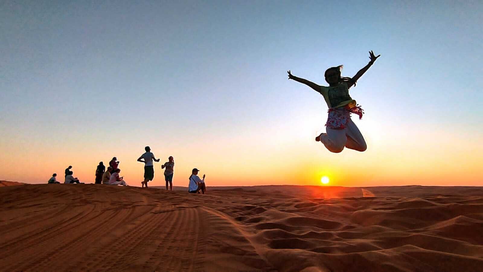 A desert landscape with a sunset, people sitting and standing on sand dunes, and a person jumping in the air.