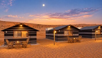 Paisaje desértico con tres cabañas, cada una con una ventana y una mesa con sillas.