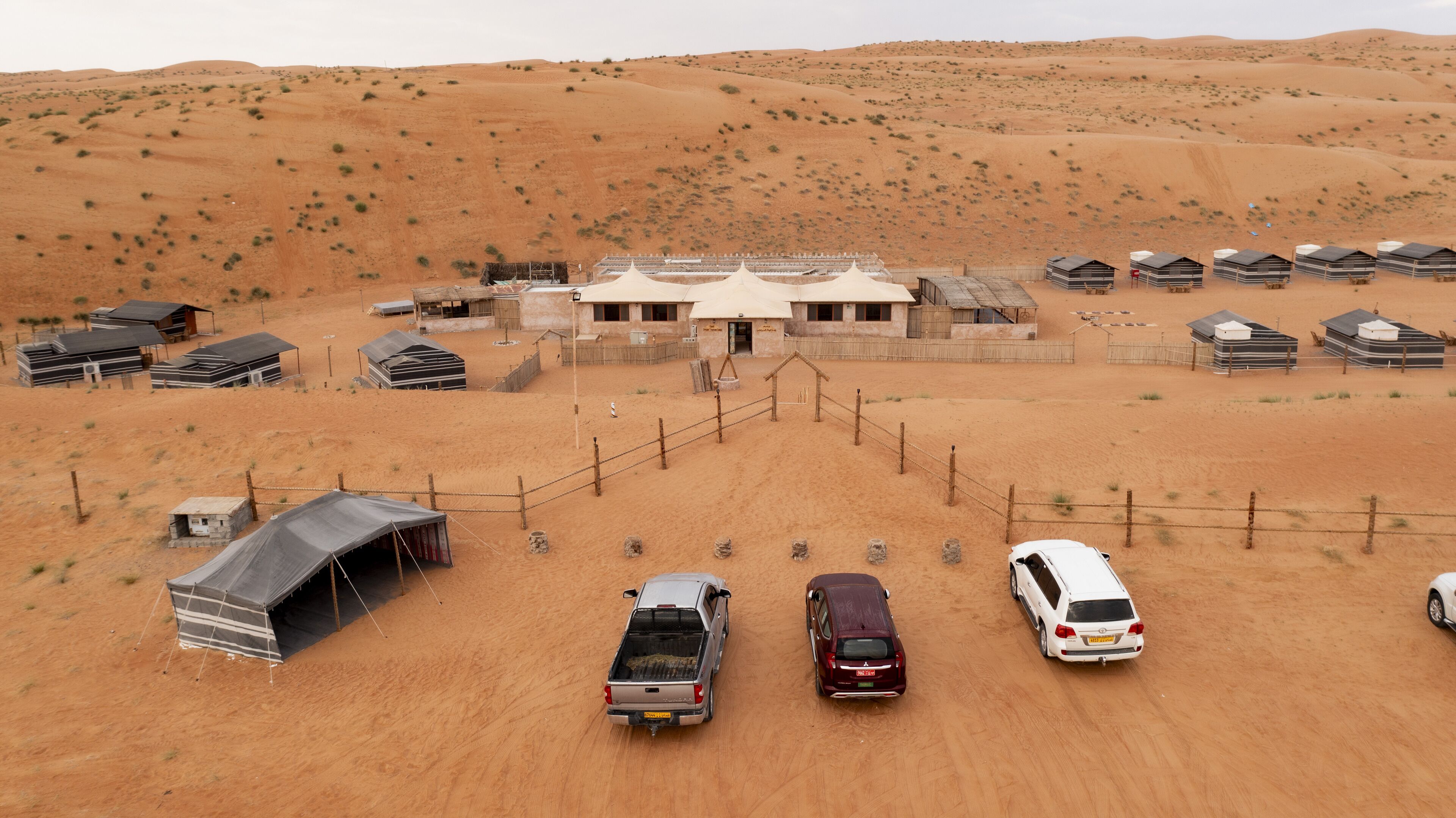 Un hôtel situé dans le désert, composé de plusieurs tentes et de chalets, avec un parking clôturé où sont garés plusieurs véhicules, le tout sur fond de dunes de sable.
