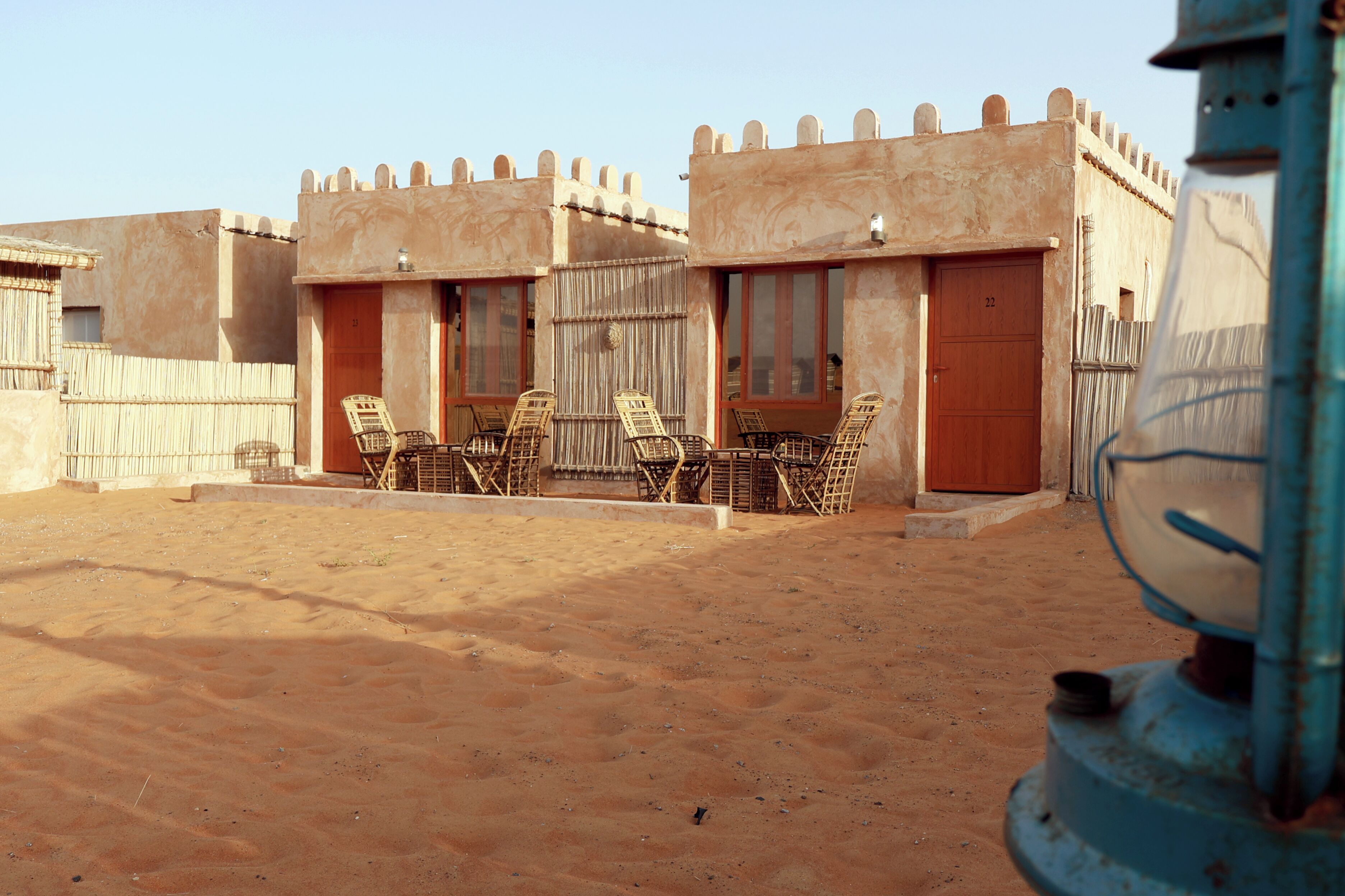 A desert-style building with multiple rooms, each featuring a wooden door and windows. There are several wicker chairs arranged outside, suggesting a seating area.