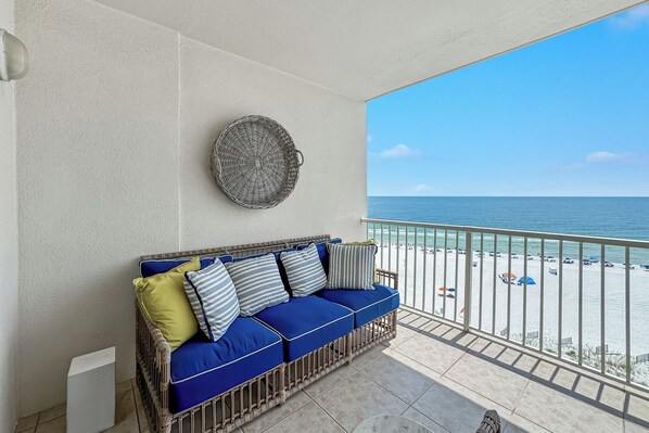 A balcony with a wicker sofa, a decorative wall piece, and a view of the beach. - Admirals Quarters 703-New-Custom Decorated-Built-in Bunks-Beachfront-Luxury-View (Orange Beach)