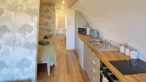 Eine kompakte, moderne Küche mit Holzausstattung, einer Mikrowelle und einem Spülbecken. Im angrenzenden Raum ist ein Bett mit einer grünen Decke zu sehen.
