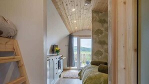 Ein schmaler Flur mit einer Holzlade­leiter links, einem Bett mit grüner Decke rechts und einer Küchenzeile mit Schränken und Spüle.