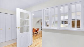 Ein Zimmer mit Holzboden, weißen Wänden und einer weißen Tür mit Glasscheiben. Im Hintergrund ist eine Essecke mit orangefarbenen Stühlen und einem Tisch zu sehen.