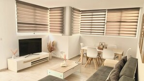 Una sala de estar moderna con un televisor de pantalla plana, un sofá, una mesa de comedor con sillas y ventanas grandes con persianas.